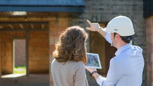 Mann mit Bauhelm zeigt auf ein Tablet, während eine Frau in einem Baustelleninneren zuschaut.
